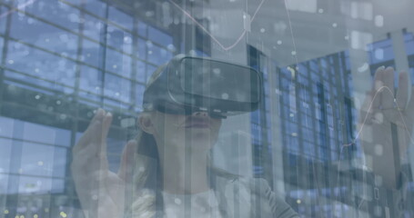 Image of financial data over caucasian woman using vr headset in office