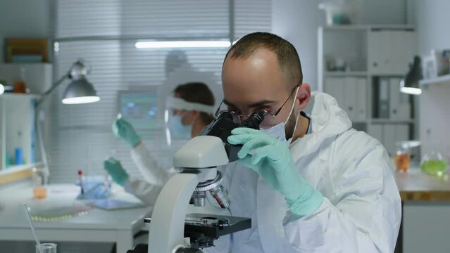 Waist up of Middle Eastern microbiologist wearing face mask, gloves and coverall working with microscope while doing research with colleagues in modern laboratory