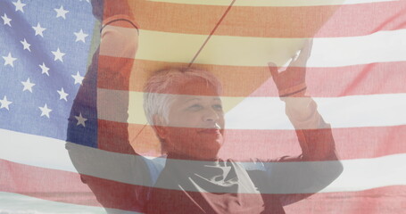Image of flag of united states of america and senior biracial woman with surfboard on beach