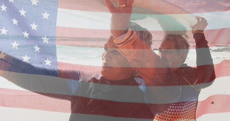Image of flag of united states of america over senior biracial couple with surfboard on beach