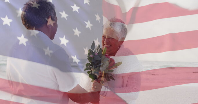 Image of flag of united states of america over senior biracial couple with flowers on beach