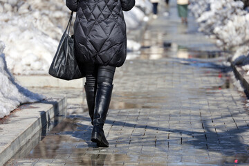 Female legs in black boots on a street with puddles. Woman walking in spring city with melting snow