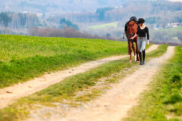 Horse and rider, saddled in riding clothes, walk side by side up a dirt road through green fields.