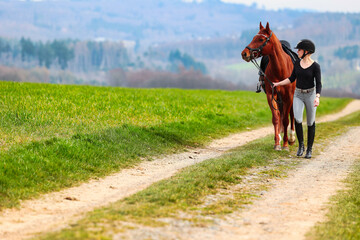 Horse and rider, saddled in riding clothes, walk side by side up a dirt road through green fields.