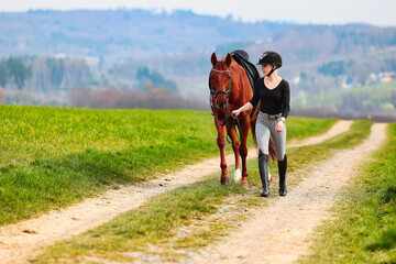 Horse and rider, saddled in riding clothes, walk side by side up a dirt road through green fields.