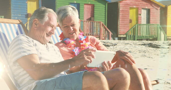 Image of glowing light over portrait of happy senior couple using tablet in deckchairs on beach