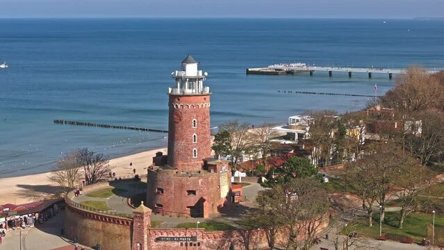 Sunny February drone footage in Kołobrzeg's port captures its lighthouse, tourists on the sandy beach, and seabirds soaring under clear blue skies.