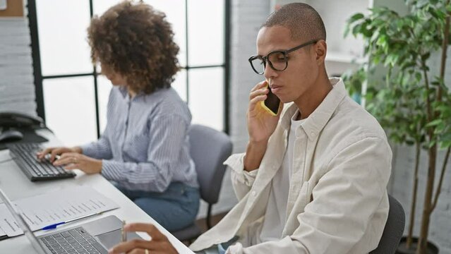 Two devoted workers - a man and woman, talking on their phones while working together on computer tasks in a professional office environment