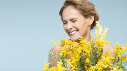 Happy portrait of confident and healthy mature woman with beautiful mimosa bouquet against blue background. Radiant smile of beautiful woman
