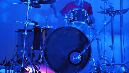 A drummer is captured in the midst of a performance on stage, surrounded by a drum kit with various cymbals and drumsticks in hand, under the bright lights of the venue.