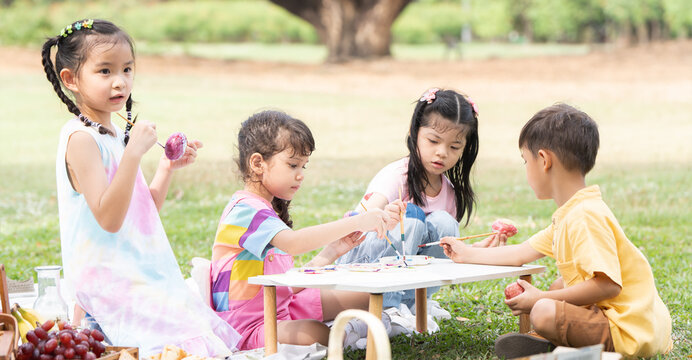 Happy group of cute little children girls painting Easter eggs together. Diverse kids holding painting brush, painting watercolor on egg while playing outdoors at park