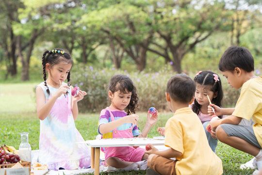 Happy group of cute little children girls painting Easter eggs together. Diverse kids holding painting brush, painting watercolor on egg while playing outdoors at park