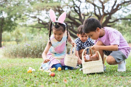 Happy group of diverse cute children hunting Easter eggs, girl wearing bunny ears. kids holding basket, picking eggs on grass while playing outdoors at park. Selective focus on little girl