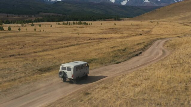 Suv car microbus driving on rural road in in autumn mountain steppe field. Picturesque wild nature snowy mountains on background. Road trip adventure travel tourism journey vacations aerial view