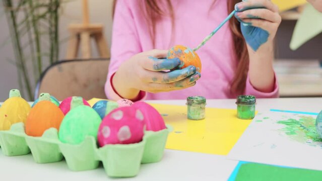 Closeup of little child painting easter eggs at home holding paintbrush coloring egg with blue and orange colors making patterns while sitting at table