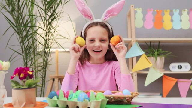 Overjoyed happy brown haired little girl wearing bunny ears headband hiding behind decorating eggs enjoying holiday preparation sitting in festive room interior