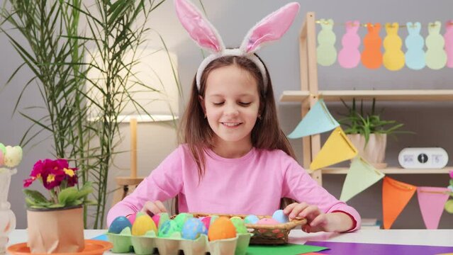 Cute charming satisfied brown haired little girl wearing bunny ears headband showing basket with many decorating eggs demonstrating her art work for holiday