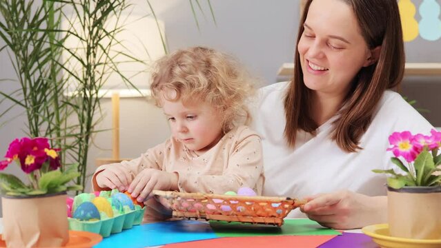 Overjoyed happy young woman with little daughter playing with Easter eggs in basket in festive home interior laughing happily while sitting together at table