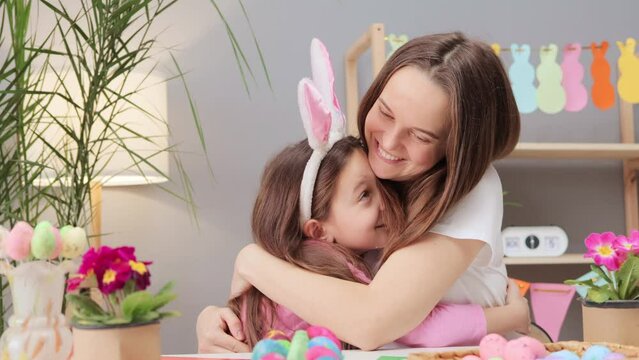 Delighted young woman with little daughter hugging each other while painting Easter eggs in festive home interior sitting at table enjoying holiday preparation activities