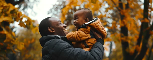 Foto op Canvas dark-skinned dad holds his little son in his arms and both laugh merrily against the backdrop of an autumn landscape close-up © Tetiana