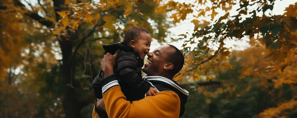 Foto op Canvas dark-skinned dad holds his little son in his arms and both laugh merrily against the backdrop of an autumn landscape  © Tetiana