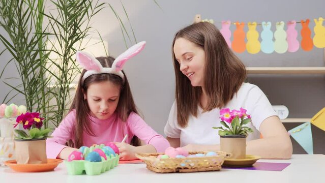 Little girl with mother sitting at table in festive home interior in kind family atmosphere drawing greeting cards and painting eggs for Easter celebration