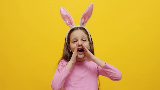 Scared hysterical small girl wearing pink bunny ears headband standing isolated over yellow background looking at camera with fear covering mouth with hands
