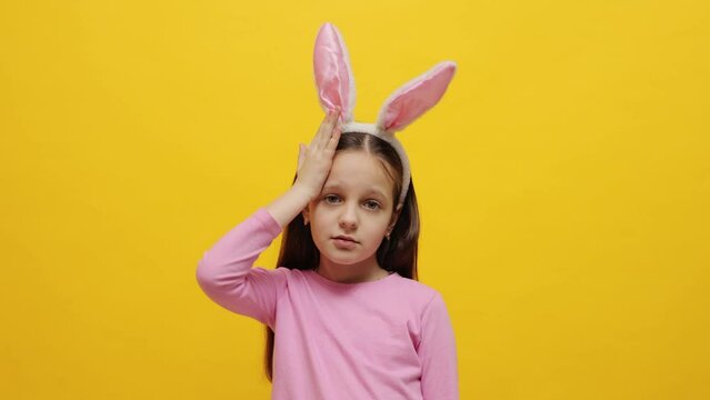 Shocked sad upset small girl wearing pink bunny ears headband standing isolated over yellow background forgot something important showing facepalm gesture
