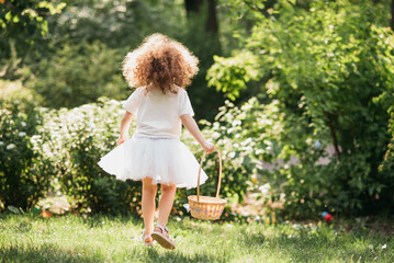 Easter egg hunt. Girl child Wearing Bunny Ears Running To Pick Up Egg In Garden. Easter tradition. Baby with basket full of colorful eggs. - 757831811