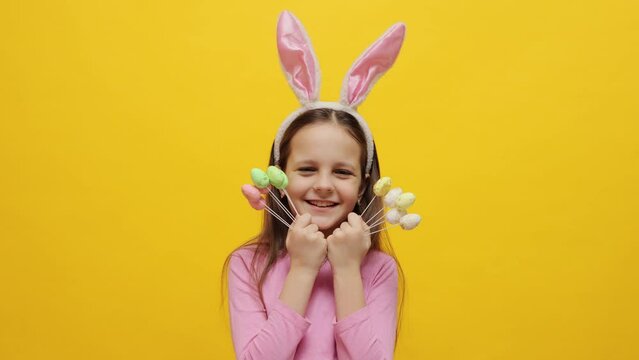 Rabbit-themed Easter party for kids. Delighted adorable little girl wearing pink rabbit ears headband holding pop cakes posing isolated over yellow background covering eyes with desserts and smiling