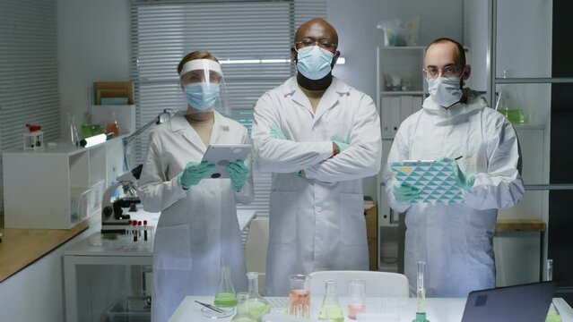 Medium slowmo portrait of multiethnic group of three scientists in white lab coats and face masks posing for camera in modern laboratory