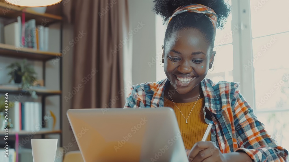 Poster the portrait of a smiling woman at her desk while she uses a laptop and writes in a notebook, taking