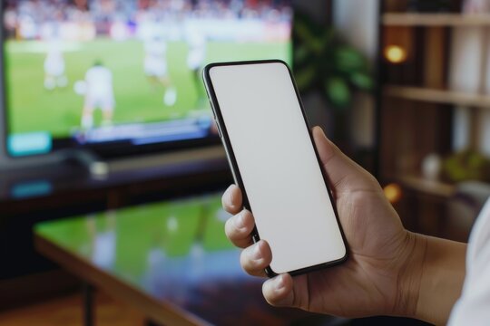A person is holding a cell phone in front of a television. The television is showing a soccer game