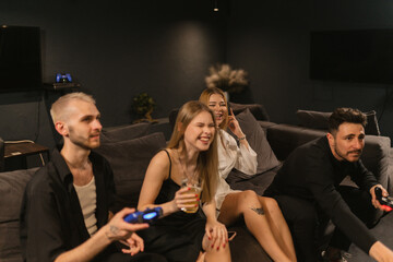 Young women laugh sitting between men playing console. Females share jovial banter with male...