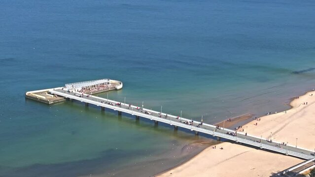 Drone footage captures Kołobrzeg pier, tourists on sandy beach, blue sky, and seabirds on a sunny day in February.