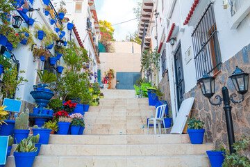 Fototapeta na wymiar Narrow street with steps, white houses and blue potted plants in ancient neighborhood Santa Cruz in Alicante old town on hillside. Costa Blanca on Mediterranean sea coast, Spain