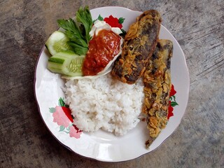 Lele krispi or crispy catfish. The catfish is seasoned and then fried in a flour coating. Served with rice, chili sauce and fresh vegetables. Top view.