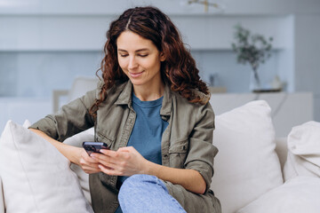 A woman sits relaxed on a sofa, deeply engrossed in her smartphone, epitomizing casual leisure in a...