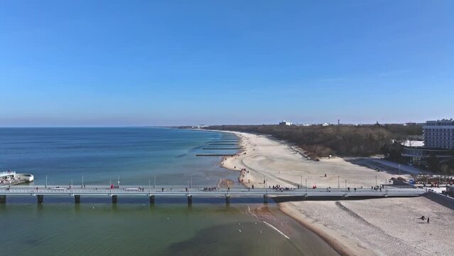 Drone footage captures Kołobrzeg pier, tourists on sandy beach, blue sky, and seabirds on a sunny day in February.