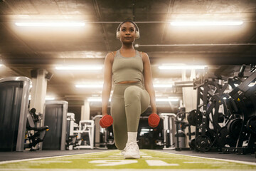 Front view of a dedicated black sportswoman doing lunges with dumbbells at gym and listening music.