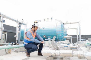 Asian male engineer working using laptop computer checks hot water pipes system or hot water boiler system tank at rooftop of building. Asian male plumber worker maintenance hot water buffer tank - obrazy, fototapety, plakaty