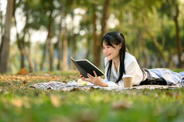 Cheerful young woman in casual clothes relaxing on green lawn in the park reading book