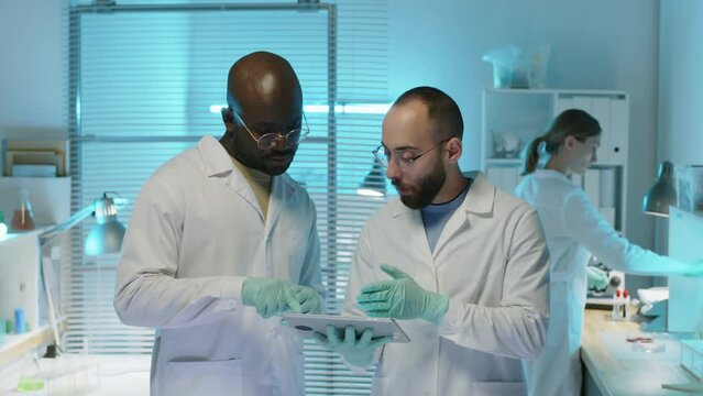 Medium shot of two ethnically diverse male scientists wearing lab coats and disposable gloves looking at digital tablet and having conversation while co-working on research in lab