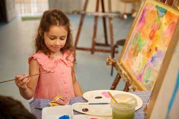 Talented little child girl artist standing in front of easel, holding brush and palette, working in fine art studio