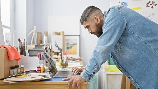 A focused bearded man using laptop in a bright art studio surrounded by painting supplies.