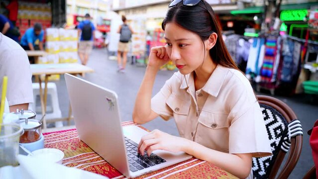 Young adult Asian woman working remotely on table in the street food stall shop in Bangkok, Thailand. Solo digital nomad working on laptop while traveling. Online freelance and business concept