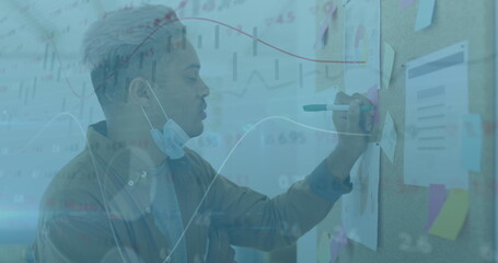 Image of financial data processing over man with face mask writing on memo note