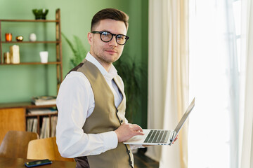 Confident man in glasses and a vest standing and working on a laptop in a well-lit room.