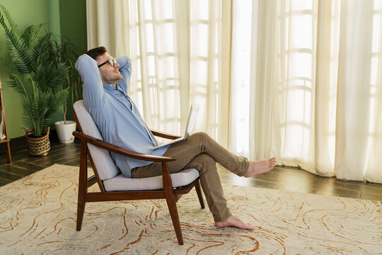 Relaxed man in a blue shirt reclines in a chair, taking a break from work, with a laptop in his lap.