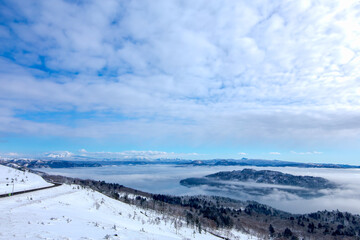 Fototapeta na wymiar 雪に覆われた山の斜面と眼下の湖の雄大な冬景色。雲の空の朝陽。北海道の美幌峠から見下ろす屈斜路湖。
